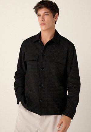 Camisetă neagră texturată cu închidere prin butoni, două buzunare la piept și mâneci lungi. Fabricată dintr-un material moale cu un model subtil.