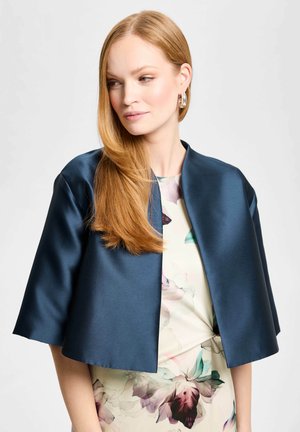 Femme aux longs cheveux roux portant des boucles d'oreilles créoles en argent, une veste courte bleu foncé et une robe fleurie, regardant de côté sur un fond uni.