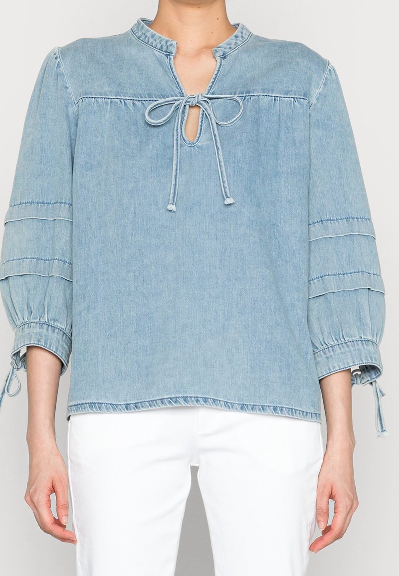 Ljusblå denimblus med en nyckelhålsutskärning och knytning, med rynkade ärmar och elastiska muddarna, samt sömdekorationer.