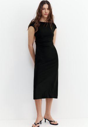 Femme aux longs cheveux bruns portant une robe mi-longue noire cintrée et des sandales à talons noires, debout contre un mur blanc.