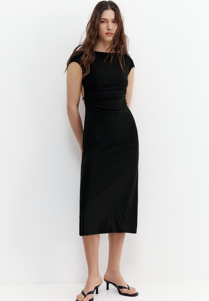 Femme aux longs cheveux bruns portant une robe mi-longue noire cintrée et des sandales à talons noires, debout contre un mur blanc.