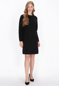 Vestido de punto negro con mangas largas y cuello redondeado. Presenta una superficie ligeramente texturizada y llega hasta la mitad del muslo. Combinado con tacones negros.