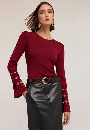 Donna che indossa una maglietta rossa a maniche lunghe aderente con ritagli decorativi sulle maniche, orecchini d'oro, gonna di pelle nera e cintura rossa con fibbia d'oro.