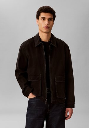 Ung mand med kort krøllet hår iført en sort lynlåstrøje med store forlommer og mørke jeans, stående mod en ensfarvet grå baggrund.
