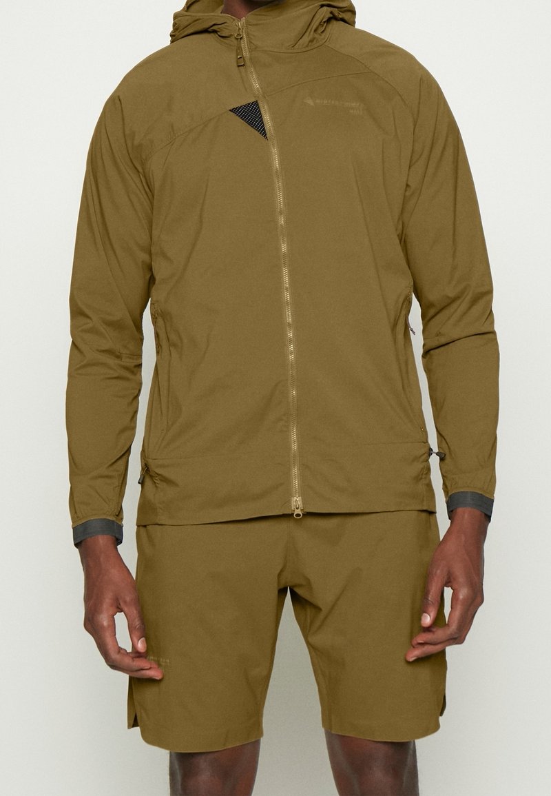 Homme portant une veste légère à capuche zippée et un short assortis de couleur vert olive, debout avec les bras détendus le long du corps.