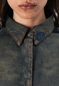 Cămașă din denim cu un model texturat, cu nuanțe de albastru și maro estompat. Prezintă o închidere cu buton, un guler în contrast și un accent de logo albastru pe guler.