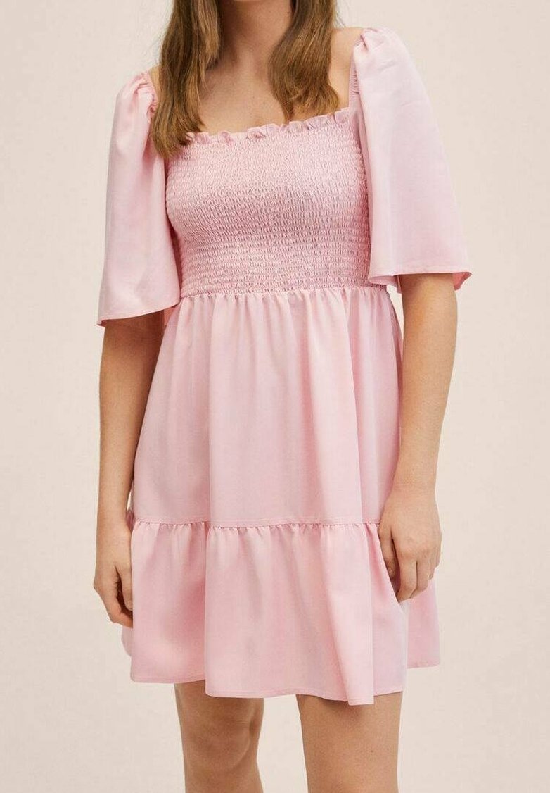 Femme portant une robe rose clair à manches courtes, avec un corsage smocké et un décolleté carré, debout devant un fond uni.