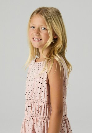 Jeune fille souriante aux longs cheveux blonds portant une robe sans manches rose clair à pois noirs, debout devant un fond uni.