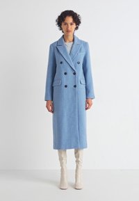 Casaco azul claro, de duplo peito, feito de um tecido macio. Apresenta dois bolsos frontais e botões pretos. Design longo e reto com uma lapela clássica.