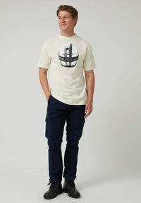 Camiseta de algodón color crema con estampado gráfico, combinada con pantalones cargo azul marino oscuro. Botas con cordones. Modelo de pie de forma casual sonriendo.