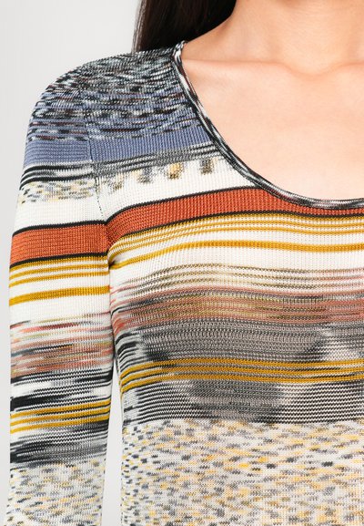 Donna che indossa un maglione a righe multicolori con scollatura rotonda, caratterizzato da tonalità di arancione, giallo, grigio, nero e bianco.