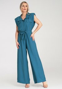 Turkusowy kombinezon z zapięciem na guziki, krótkimi rękawami, kieszeniami na piersi i dopasowanym paskiem. Szerokie nogawki zwiększają swobodę ruchów i komfort.
