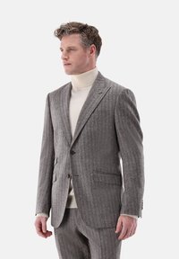 Graue strukturierte Sakkojacke mit dezenten Nadelstreifen, zwei vorderen Taschen und einem einzigen Knopfverschluss. Über einem cremefarbenen Rollkragenpullover getragen.