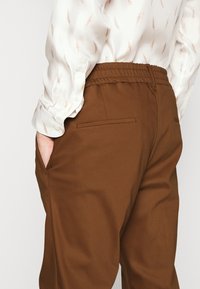 Osoba ubrana w brązowe spodnie z elastycznym pasem, z rękami w kieszeniach i w białej koszuli w jasnobrązowe wzory przypominające kłosy pszenicy.