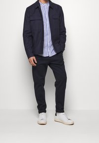 Granatowa koszulowa kurtka z dwoma przednimi kieszeniami na niebiesko-białej, paskowanej koszuli na guziki. Do tego ciemne spodnie i białe trampki.