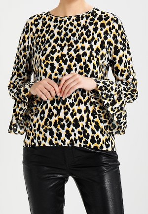 Žena v dlouhém leopardím topu s rozšířenými manžetami a černých kožených kalhotách, ruce sepnuté v úrovni pasu.