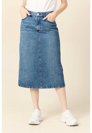 Kvinde iført en blå denim midi nederdel med lommer, kombineret med hvide sneakers og en hvid t-shirt stoppet ind, stående med hænderne i lommerne.