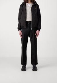 Zwart jack met ritssluiting en capuchon, grijze gebreide trui en zwarte broek. Opvallend geel logo op het jack, zwarte schoenen met een dikke zool.