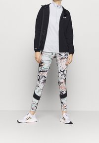 Musta huppari vaaleanharmaan vetoketjullisen paidan päällä, yhdessä värikkäiden, marmoroitujen legginssien kanssa, joissa on mustaa, valkoista ja pehmeitä pastellivärejä, sekä valkoiset lenkkarit.