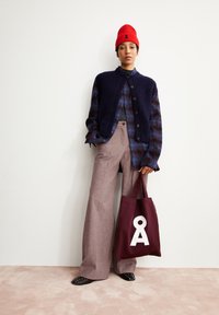 Overmaatse marineblauwe gebreide vest over een geruit shirt, gecombineerd met lichtbruine wijde broek en een bordeauxrode totebag met een witte grafiek.