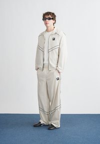 Σετ φόρμας σε ανοιχτό μπεζ χρώμα με μαύρες λεπτομέρειες, που περιλαμβάνει μπουφάν με φερμουάρ και άνετο παντελόνι. Διαθέτει λογότυπα και γυαλιστερό, υφασμάτινο ύφασμα με υφή.