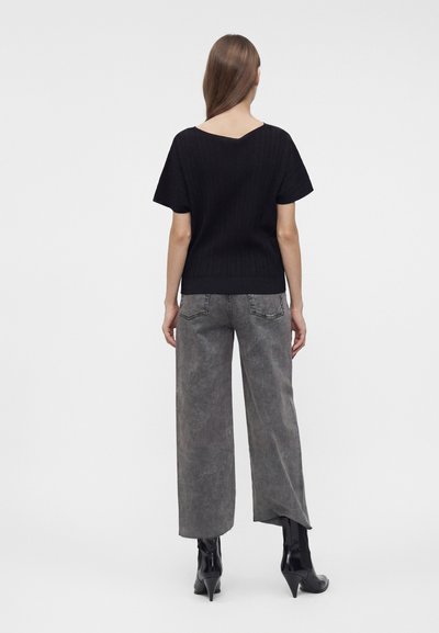 Femme aux longs cheveux bruns portant un haut noir à manches courtes, un jean gris à jambes larges et des bottines noires à talons, debout de dos.