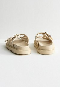 Sandalias trenzadas beige con una tira ancha y hebilla, suela de goma gruesa, planta suave y detalles texturizados en la parte superior.
