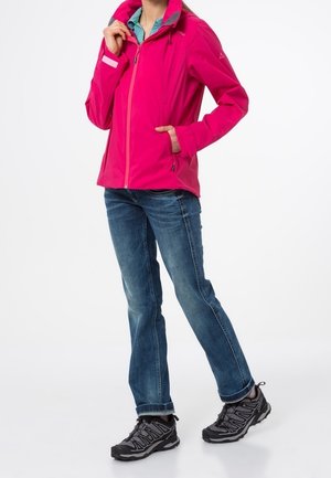 Persoon met een felroze ritsjack, blauwe jeans en zwarte wandelschoenen, staand met één hand in de zak.