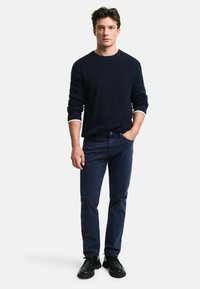 Granatowy sweter w prążki z białym wykończeniem przy mankietach, w połączeniu z ciemnymi dżinsami i czarnymi butami. Model stoi na jasnym tle.