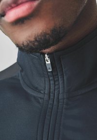 Chaqueta negra con cremallera en la parte delantera y cuello alto, con una textura suave, costuras visibles y una cremallera plateada con un detalle triangular.