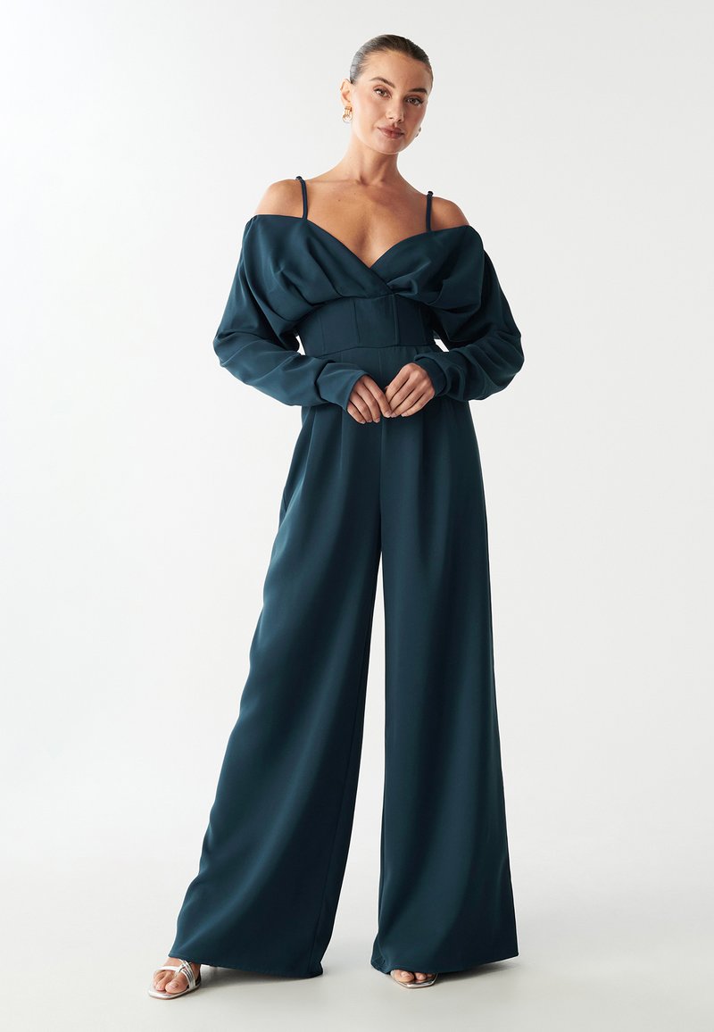 Tīrā zāles zaļa kombinezons ar plecus atklājošu dizainu, garām piedurknēm, platām kājām un pieguļošu jostasvietu. Izgatavots no gludas auduma ar minimālām rotājumiem.