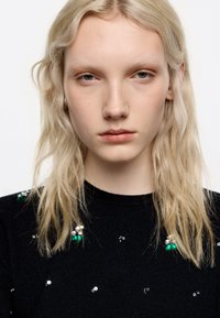 Suéter negro con cuello redondo, adornado con cuentas decorativas en plateado y verde, y con una textura suave. El cabello es ondulado y está parcialmente visible.
