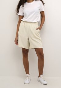 Shorts beiges texturés avec une taille élastique et des poches latérales, associés à un T-shirt blanc uni et des baskets blanches.