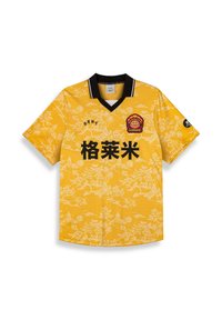 Jersey amarillo de manga corta con cuello negro. Presenta un patrón floral en blanco y texto negro. Incluye un logo rojo en el lado izquierdo del pecho.