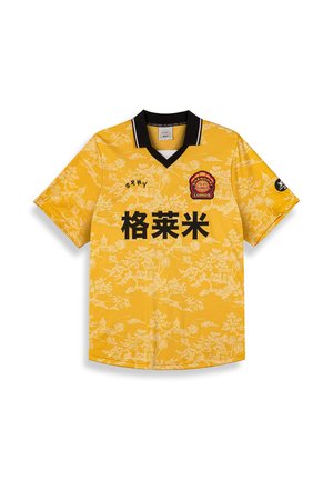 Jersey amarillo de manga corta con cuello negro. Presenta un patrón floral en blanco y texto negro. Incluye un logo rojo en el lado izquierdo del pecho.