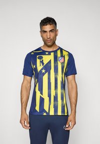 Blå sports t-shirt med gule grafiske striber og mønstre. Har et logo på brystet. Korte ærmer og rund hals.