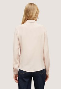 Blusa rosa chiaro con maniche lunghe, colletto e texture liscia. Presenta un orlo arrotondato e polsini con bottoni, abbinata a jeans scuri.
