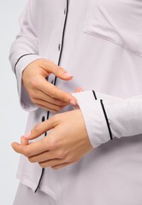 Cămașă butonată de culoare violet deschis, cu margini negre și buzunar. Materialul pare moale și neted; modelul își ajustează lungimea mânecilor.