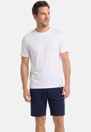 Man draagt een effen wit T-shirt met korte mouwen en marineblauwe shorts, staat en kijkt naar de zijkant tegen een effen achtergrond.
