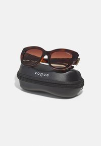 Tortoiseshell sunglasses with brown gradient lenses sit on a black, zippered case featuring white "vogue" branding.
