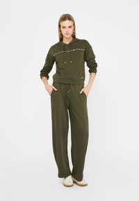 Donna in felpa verde oliva e pantaloni larghi abbinati, con sneakers bianche, in piedi con le mani nelle tasche su sfondo bianco.