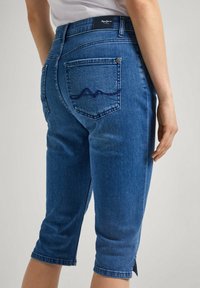 Džínové capri kalhoty středně modré barvy s designem pěti kapes. Mají prošité zadní kapsy a úzký střih, končící pod koleny.