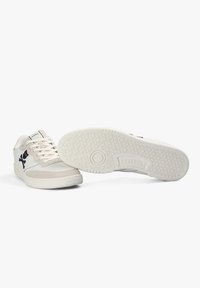 Zapatillas blancas de caña baja con logotipo negro en el lateral, acentos de ante beige, cordones blancos y suela de goma texturizada que muestra el nombre de la marca "Scalpers."