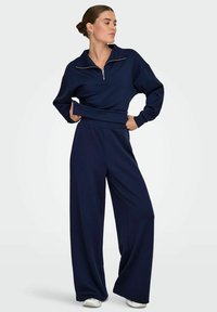 Pull-over bleu marine avec une fermeture éclair quart et des poignets côtelés, associé à un pantalon large en matériau similaire, créant un look athlétique cohérent.