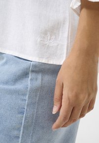 Camicia bianca in lino con un delicato ricamo di fiori di loto vicino all'orlo; abbinata a jeans azzurri, che mostrano una trama morbida.