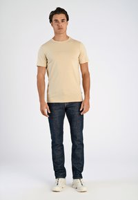 Beige värinen lyhythihainen paita hienovaraisella brändäyksellä, yhdistetty tummansinisiin farkkuihin ja valkoisiin lenkkareihin. Yksinkertainen muotoilu, sileä pintakuvio, istuva leikkaus.