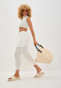 Crop top en maille blanche et jupe longue assortie avec un motif découpé, accompagnés d'un sac à main beige arrondi et de sandales à enfiler de couleur claire.
