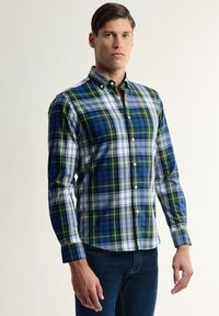 Camisa de cuadros en azul, verde y blanco. Presenta un cuello extendido y mangas largas. Está hecha de un material de algodón ligero.