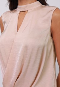 Top sin mangas color rosa claro con cuello alto y abertura en forma de llave; hecho de una tela suave y brillante con un frente drapeado.