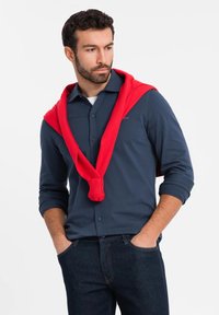 Marineblauwe longsleeve shirt met een rode trui om de nek, gedragen met donkere spijkerbroeken. Glad materiaal met knoopsluiting en merkdetail.